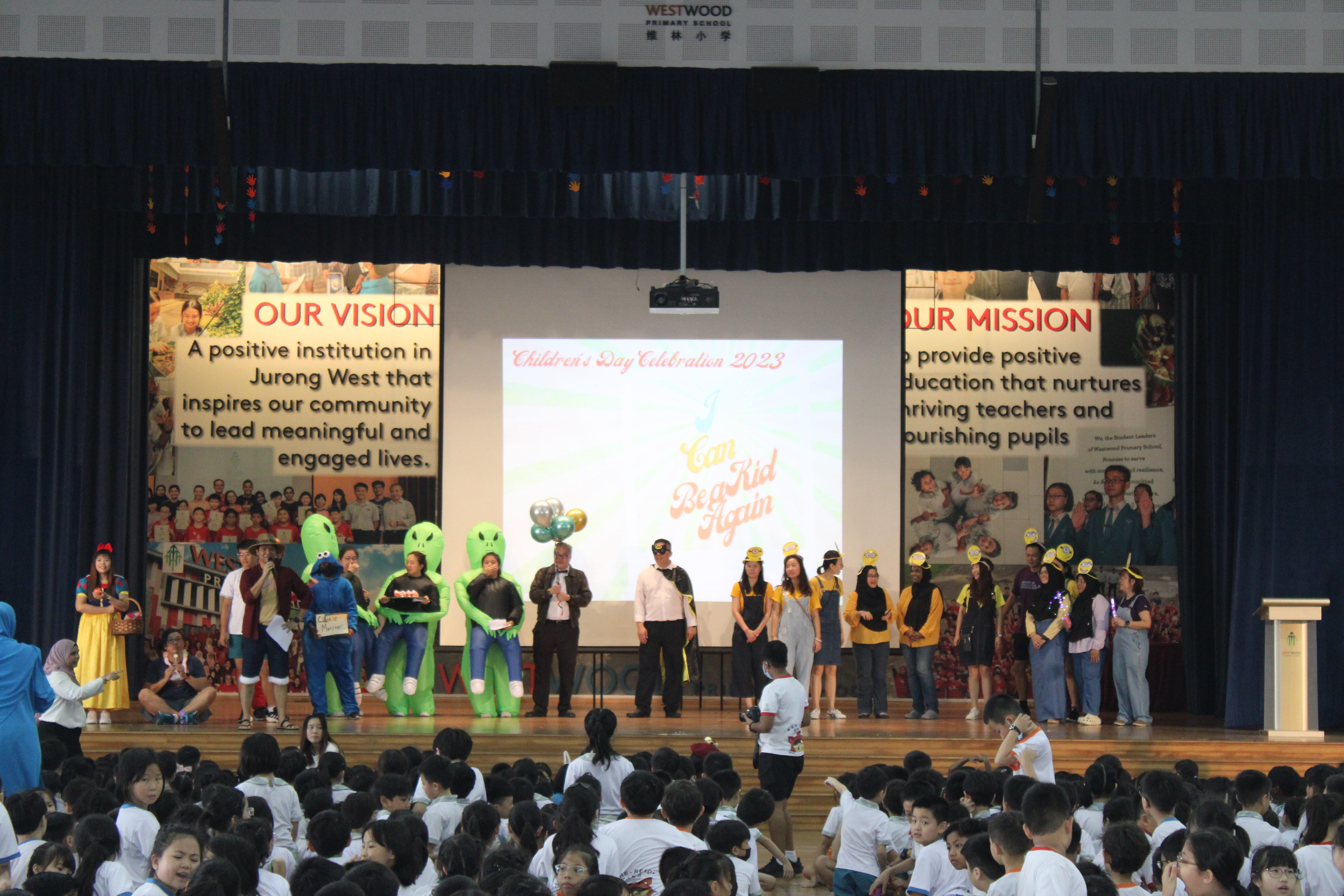 School auditorium stage with costumed performers and banners, audience of children seated in front